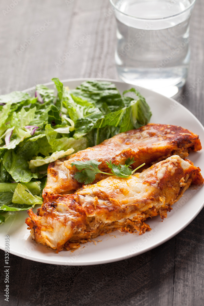 Spicy chicken enchiladas with a side of light green salad on a dark wood background vertical shot Spicy chicken enchiladas with a side of light green salad on a dark wood background vertical shot