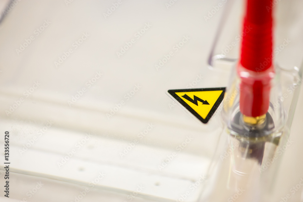 Close-up macro detail of a triangle electrical warning sign on the lid of an electrophoresis chamber, next to a red terminal wire. Cancer research and healthcare.
