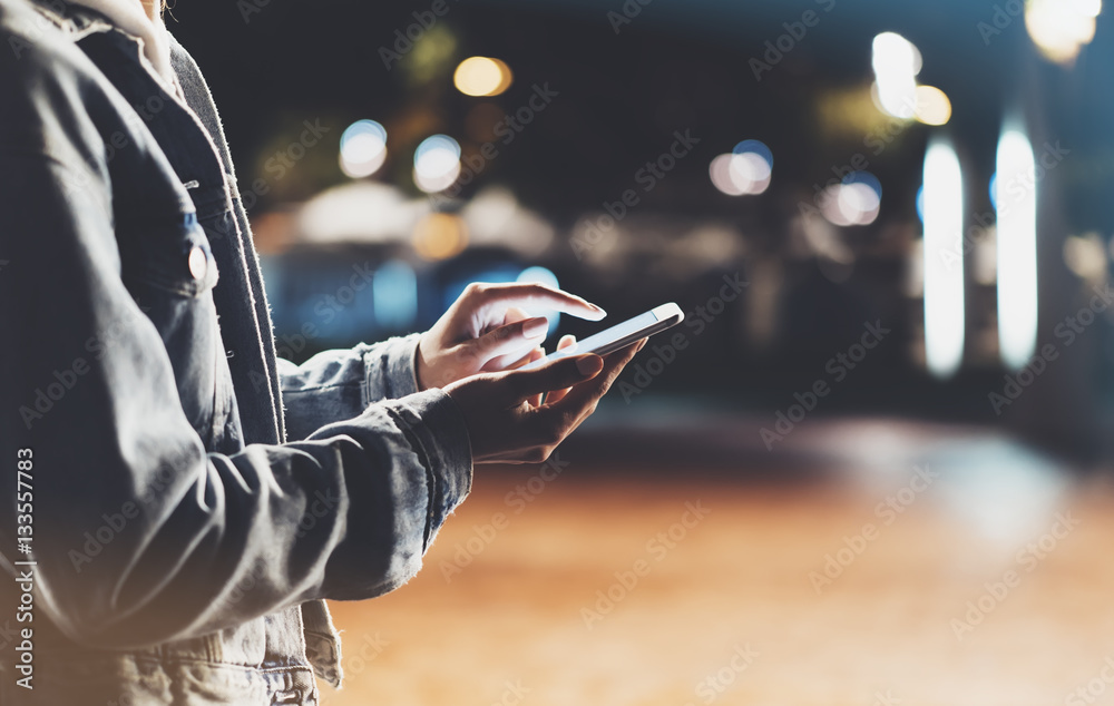 Girl pointing finger on screen smartphone on background illumination bokeh color light in night city, hipster using in hands and texting mobile phone, mockup glitter street, content lifestyle