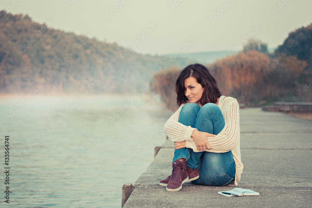 Young girl trying to warm up herself. Sitting by lake. Cold spring or autumn morning. 