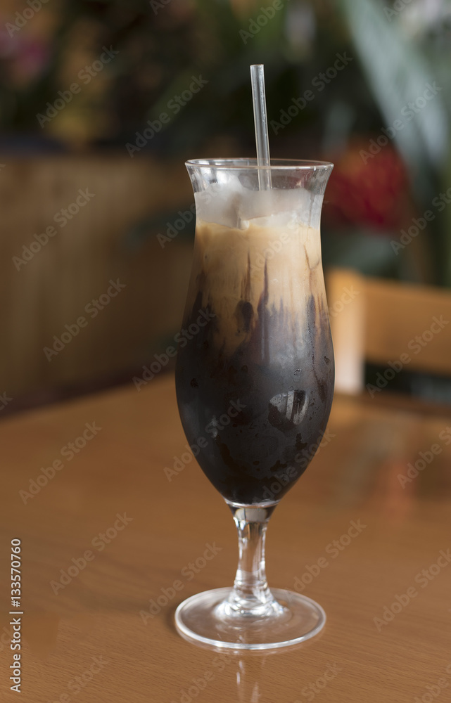Tall shapely glass of Thai iced coffee with a straw on a brown wood table against a blurred restaurant background Tall shapely glass of Thai iced coffee with a straw on a brown wood table against a blurred restaurant background