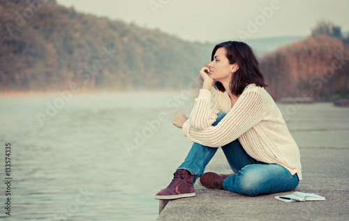 Thoughtful woman sitting by lake. Enjoying the early morning. 