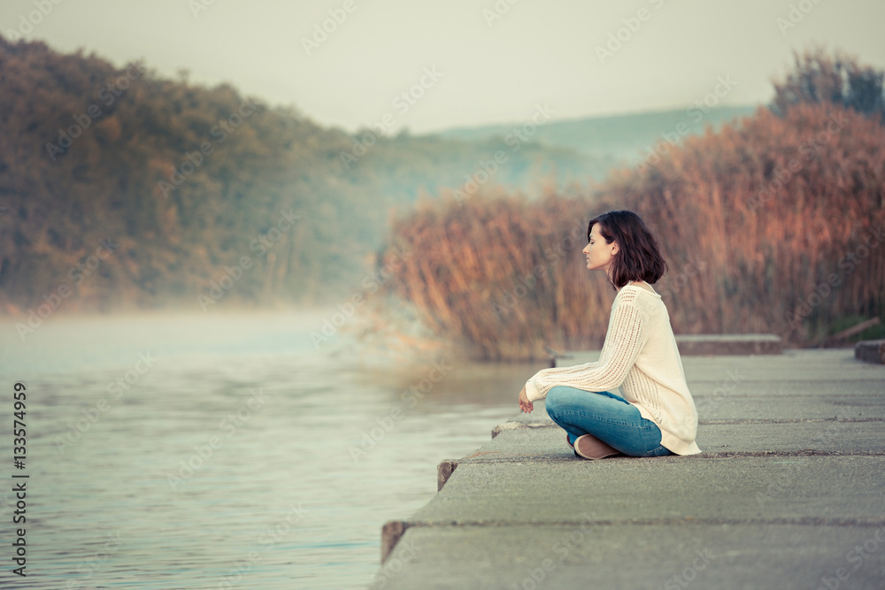 Relaxation by lake. Young woman sitting and looking away. Chilly morning. Relaxation by lake. Young woman sitting and looking away. Chilly morning.
