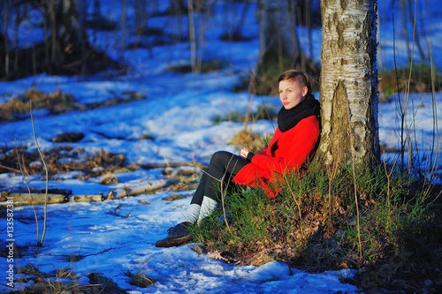 Fototapeta Naklejka Na Ścianę i Meble -  young smiling girl in red coat sitting on the thawed patch with green grass near a tree in the snow in a fabulous wild forest.
