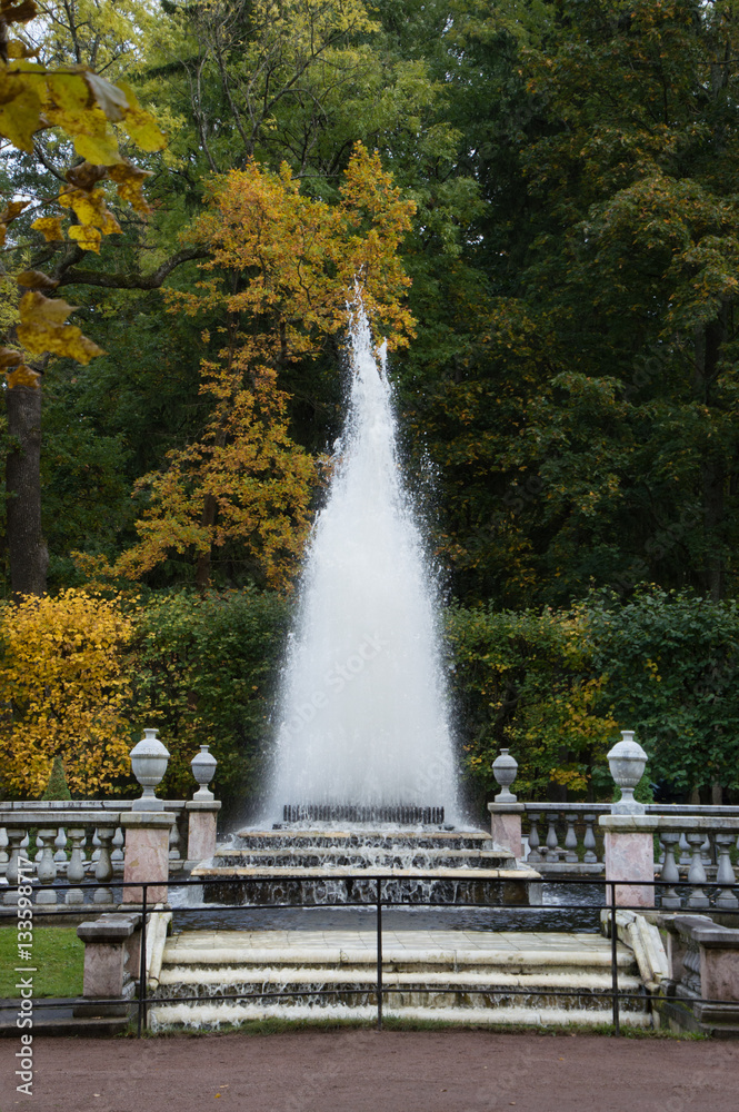 Naklejka premium Pyramid Fountain at Peterhof with dirt path and stone steps leading to the fountain, stone banisters around the fountain and fall foliage in the background.