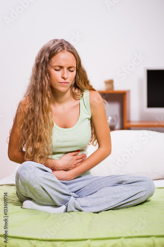 Young woman is sitting in her bed with stomachache.