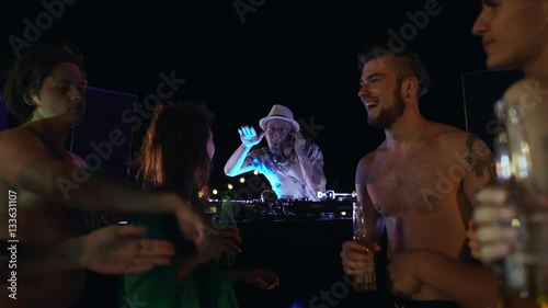 DJ in hat and glasses playing music at night pool party while topless males and female dancing laughing smiling drinking beer and cocktails in slowmotion