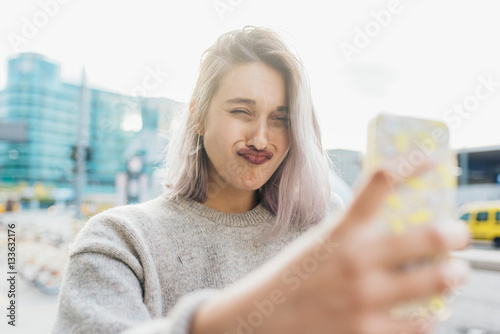 Young beautiful caucasian purple grey hair woman outdoor in the city outdoor in city back light taking selfie with smart phone hand hold doing grimace - vanity, social network, sharing concept photo