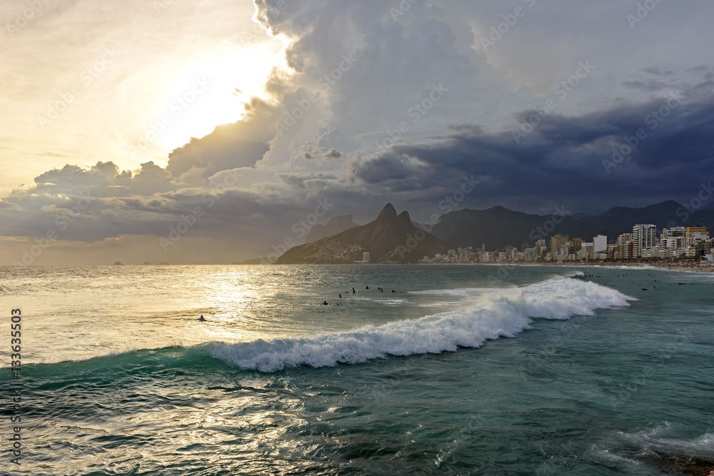 Arpoador beach in Rio de Janeiro, with its stones, buildings and the seaside during sunset and the Two Brothers hill in background