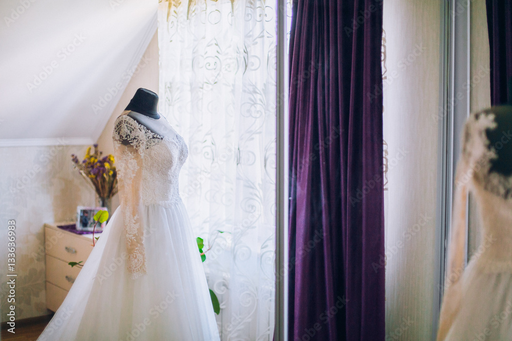 Beautiful blonde bride in robe posing near white wedding dress on mannequin Beautiful blonde bride in robe posing near white wedding dress on mannequin