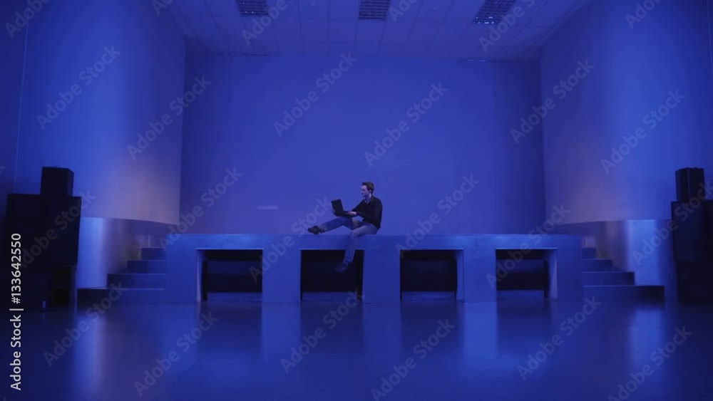 A young man sits alone on an empty dance floors and uses laptop to ...