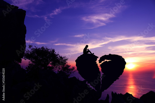 Women sit on broken heart-shaped stone on a mountain with purple sky sunset background.Silhouette Valentine background concept. 