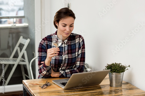 Obraz na plátně Pretty young brunette taking a sip of wine while using a compute