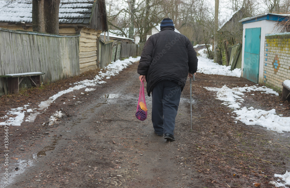 Elderly peasant with walking stick goes on an empty street of rural village Pidstavky, Sumskaya oblast, Ukraine