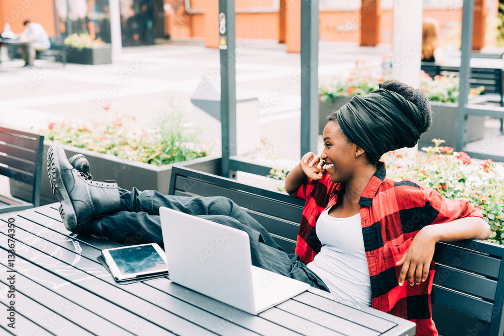 Fototapeta premium Young beautiful afro black woman sitting outdoor with legs on the table, relaxed, using computer and tablet, smiling - working, business, studying concept