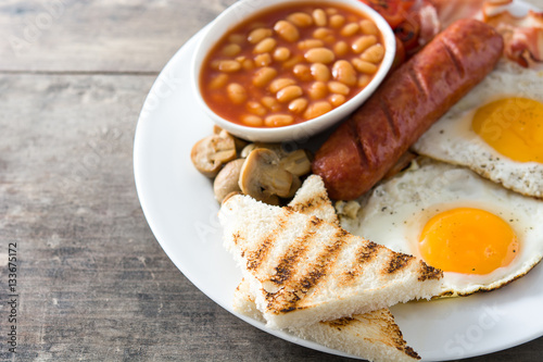 Traditional full English breakfast with fried eggs, sausages, beans, mushrooms, grilled tomatoes and bacon on wooden background
