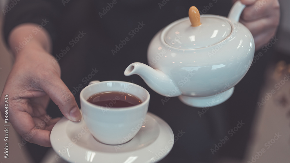 Hand holding a teapot pouring tea into a white cup on a wooden table. Hand holding a teapot pouring tea into a white cup on a wooden table.