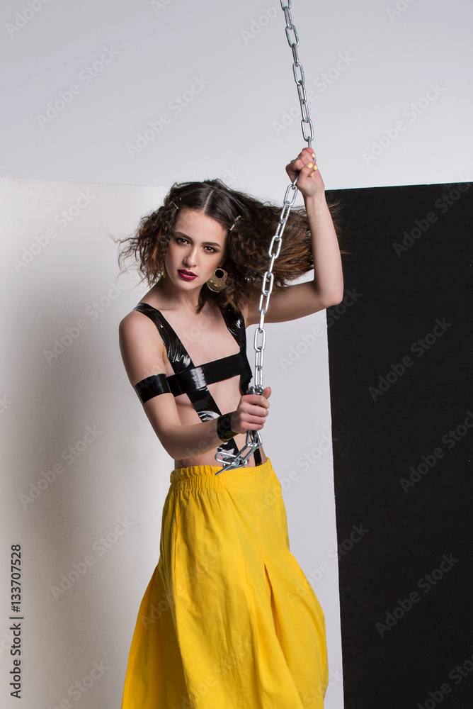 Fashion photo of beautiful girl posing in studio. Wearing yellow shorts, black boots. Straps over breast. Holding chain. Curly hairstyle.