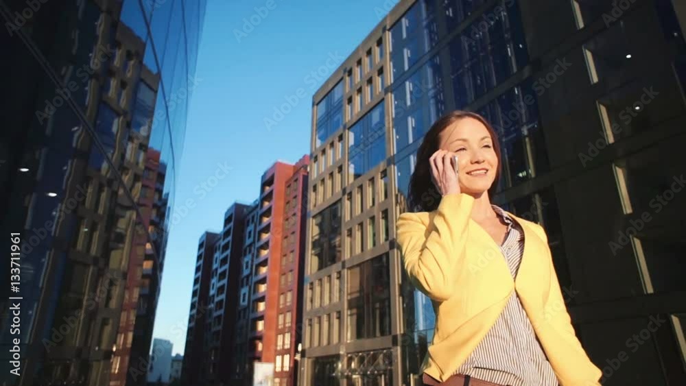 Low angle view video of Young beautiful girl in a yellow jacket near office with the phone. Business woman speaking by smartphone at the business center. Bright sunlight in the morning. Low angle view video of Young beautiful girl in a yellow jacket near office with the phone. Business woman speaking by smartphone at the business center. Bright sunlight in the morning.