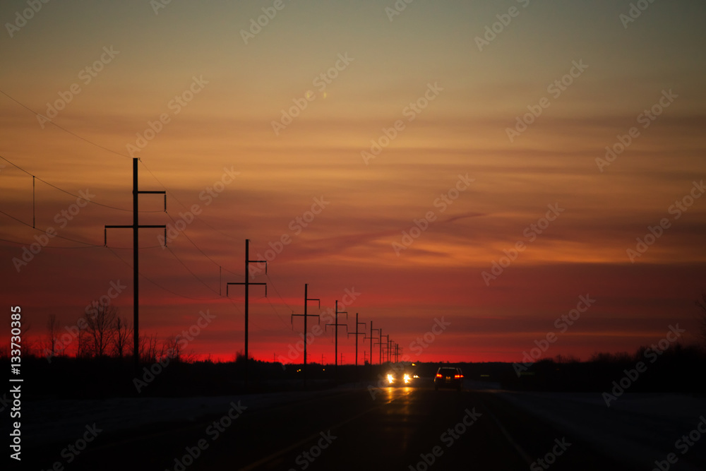 Headlights and taillights of cars on a highway at night under a colorful sunset in rural landscape