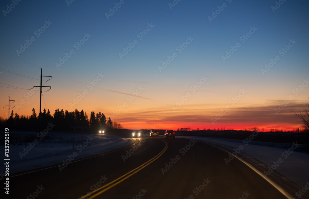 Cars travelling along a curving highway lines with power lines under colorful sunset at night in rural landscape