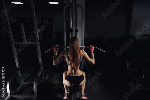 Back view. Portrait of young woman working out on exercise machine inside gym. Woman doing seated exercises at a gym.