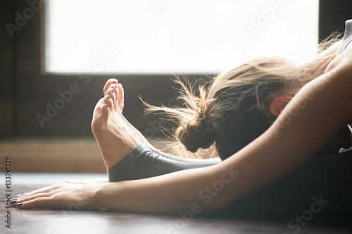 Young woman practicing yoga, sitting in Seated forward bend exercise, paschimottanasana pose, working out, wearing sportswear, grey pants, bra, indoor, home interior background, close up