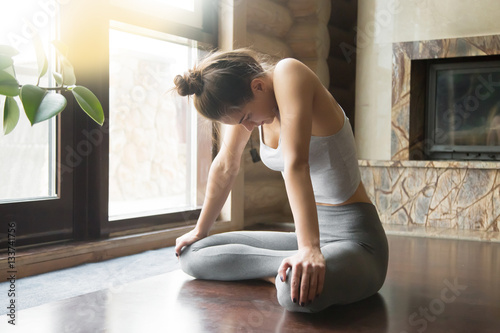 Young attractive woman practicing yoga, sitting in Uddiyana Bandha exercise, Upward Abdominal Lock pose, working out, wearing sportswear, grey pants, bra, indoor full length, home interior background