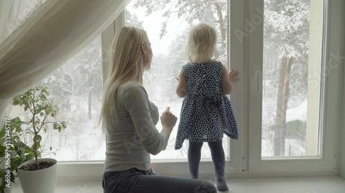 Mother is showing the winter landscape to her little daughter through a window, pointing out to something, the girl is standing  on a windowsill