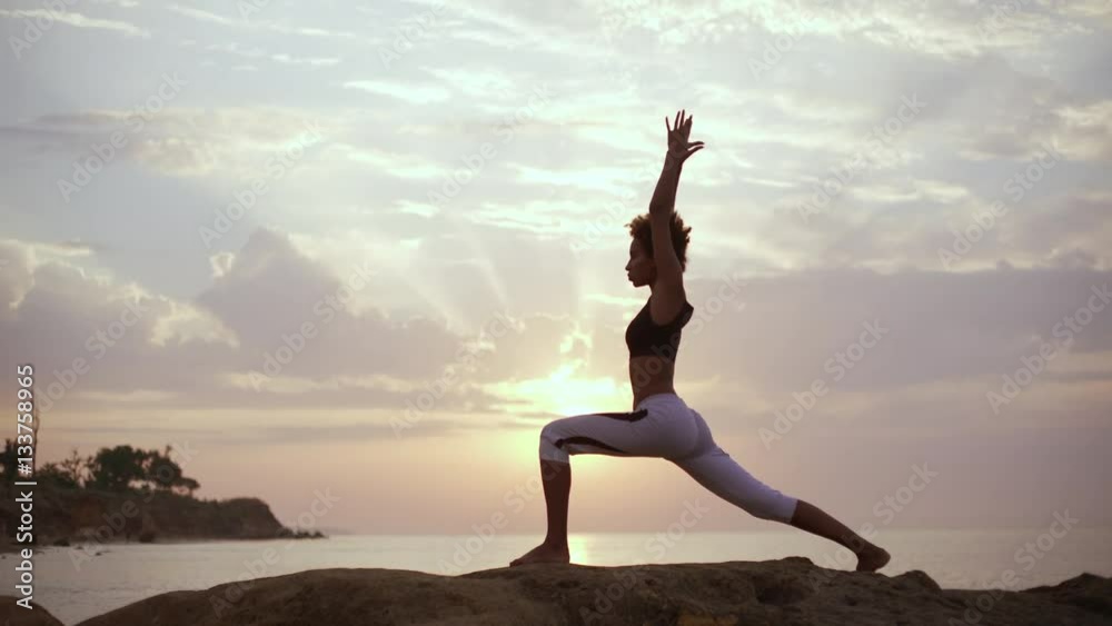 Concentrated dark-skinned woman with Afro hairstyle doing yoga exercises on rocks near sea. Silhouette Concentrated dark-skinned woman with Afro hairstyle doing yoga exercises on rocks near sea. Silhouette