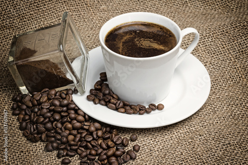 Higher angle view of the vintage photo of the white cup of coffee standing on the sackcloth. Around it are scattered coffee beans and ground coffee glass 
