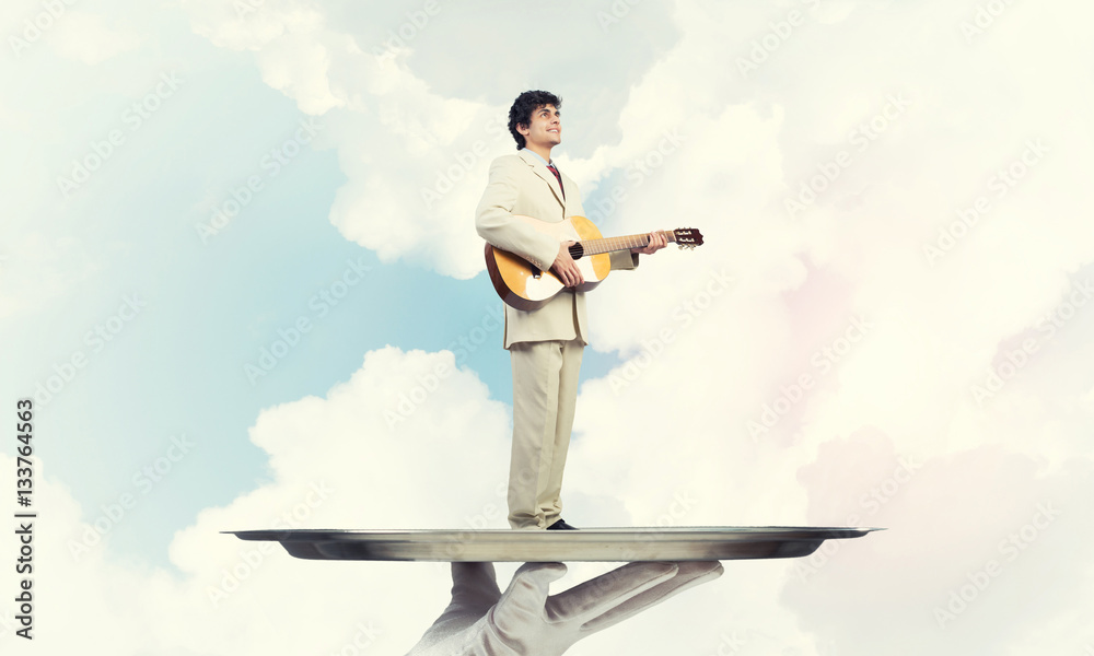 Businessman on metal tray playing acoustic guitar against blue sky background Businessman on metal tray playing acoustic guitar against blue sky background