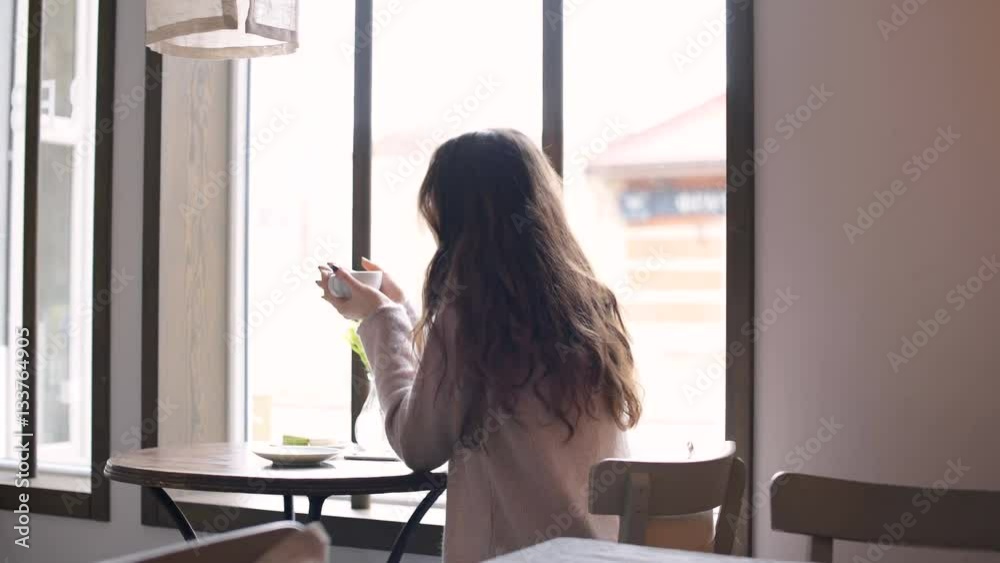 Back view of a young woman with long hair sitting and looking at the window while drinking coffee in cafe Back view of a young woman with long hair sitting and looking at the window while drinking coffee in cafe