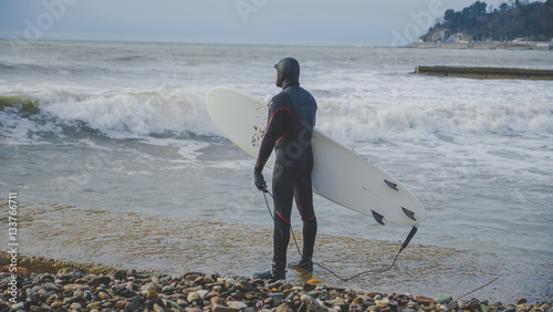 Зимний сёрфинг, сёрфер зимой в гидрокостюме идёт по пирсу в море с волнами