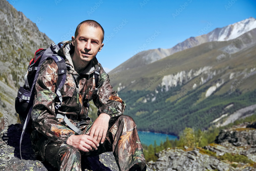 Survival in the wild. A man in camouflage resting among the mountains. Stalker, survive in the woods. Lake in the mountains. Snowy peaks, Altai
