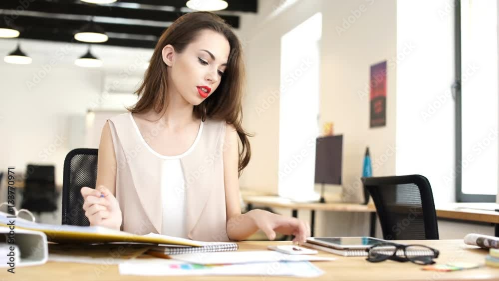 Young business woman picking up mobile phone while making notes in notebook at the table in office Young business woman picking up mobile phone while making notes in notebook at the table in office