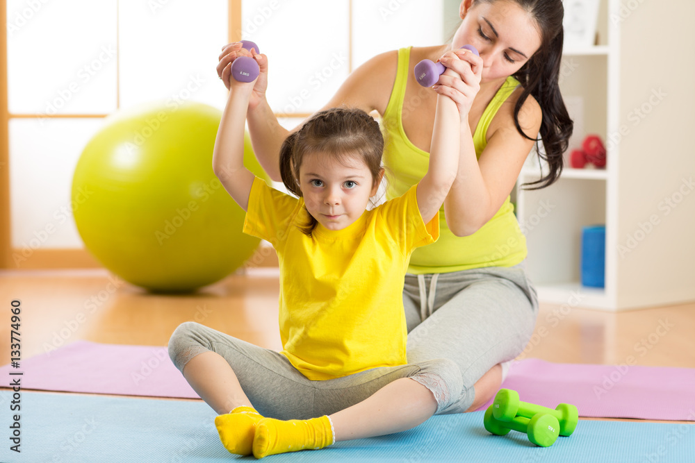 Fototapeta premium Woman helps child doing fitness exercises with dumbbells in gym