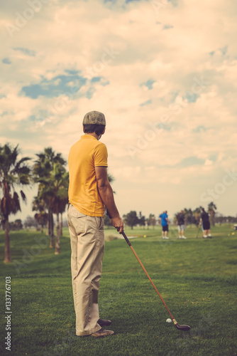 Back view of active man holding playing vintage wooden golf club on palm tree grass field outdoors background.
