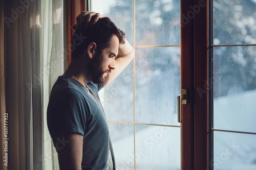 Young sad mad sitting by the window in regret photo