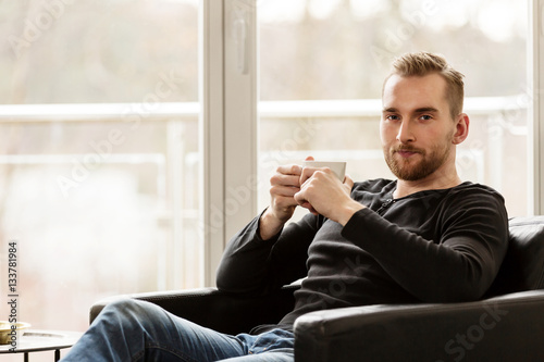 An attractive blonde man sitting down relaxing in his lounge chair holding a mug and looking at camera. Big bright large windows behind him.