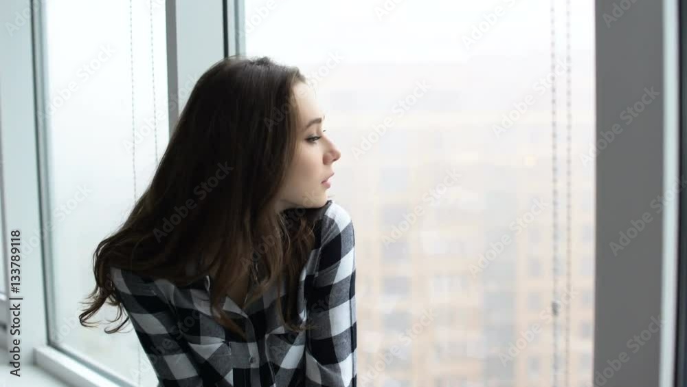 Beautiful young woman looking through window while sitting at windowsill at home