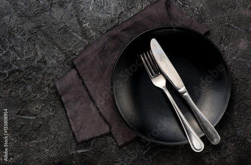 Fork, knife with napkin, roses and black plate. On dark table background.