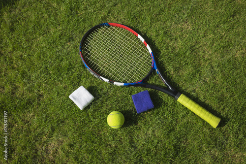 Tennis ball, racket and wristbands on grass field ground under sunlight