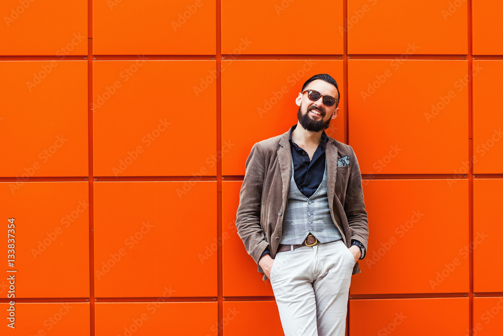 Young confident hipster man with beard in glasses posing on the orange background. Outdoor. Cute man wearing a vest, black shirt and light trousers. Man keeps hands in pockets and looking forward Young confident hipster man with beard in glasses posing on the orange background. Outdoor. Cute man wearing a vest, black shirt and light trousers. Man keeps hands in pockets and looking forward