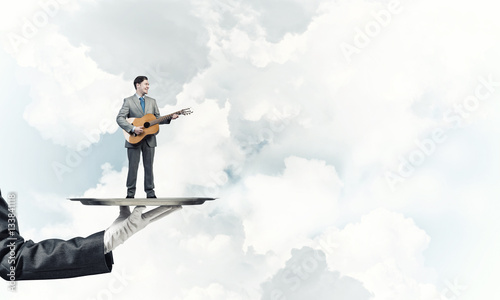 Businessman on metal tray playing acoustic guitar against blue sky background