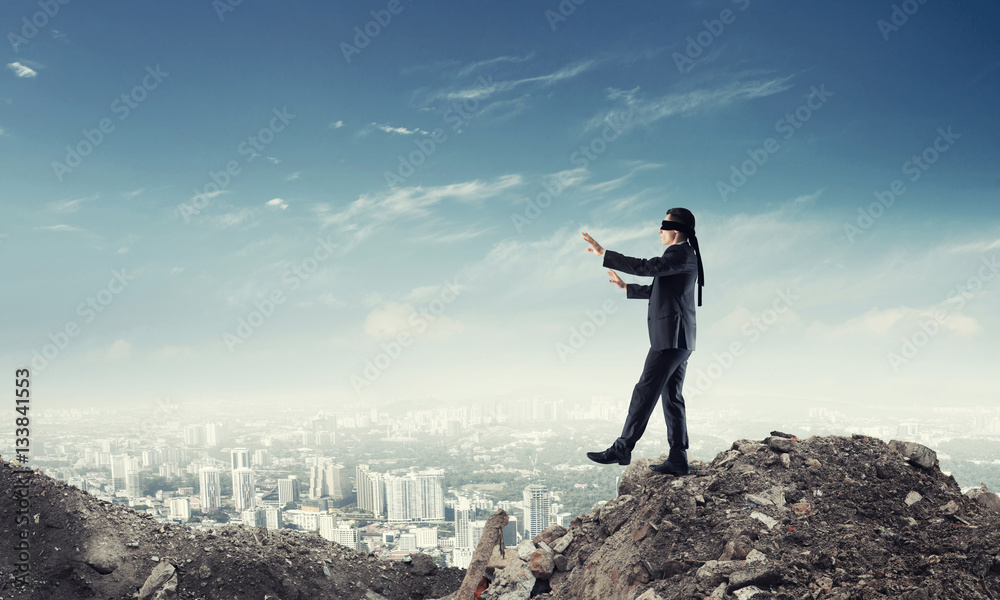 Young businessman in blindfold walking carefully and cityscape at background