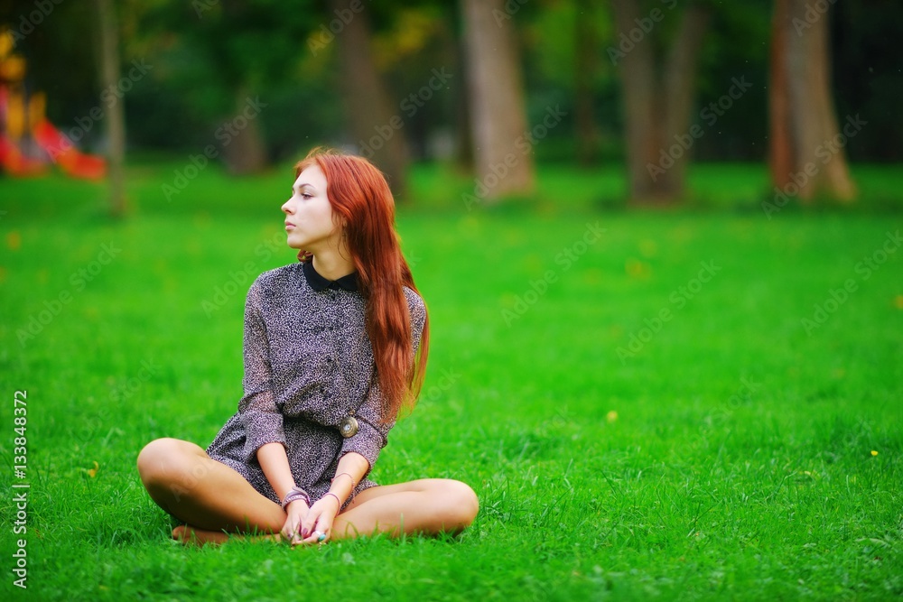 Obraz premium Portrait of young calm long-haired girls brown-haired women in short dress sitting in a Lotus position on a green lawn in a Park on a clear Sunny day.