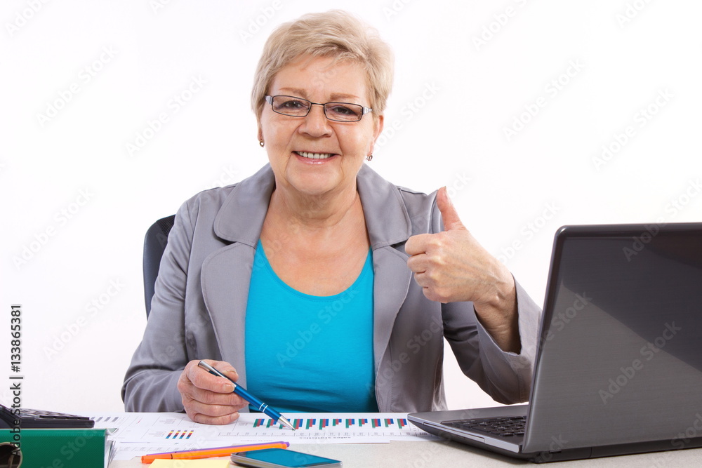 Elderly business woman showing thumbs up and working at her desk in office, business concept