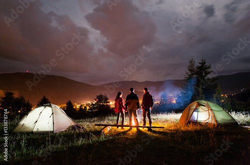 Rear view pair holding hands and their friend stands near campfire and looks at it under cloudy sky on the background mountains and luminous town in the evening