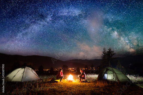 Romantic couple sitting at a campfire near tents in the night under incredibly beautiful starry sky and Milky way. Night camping
